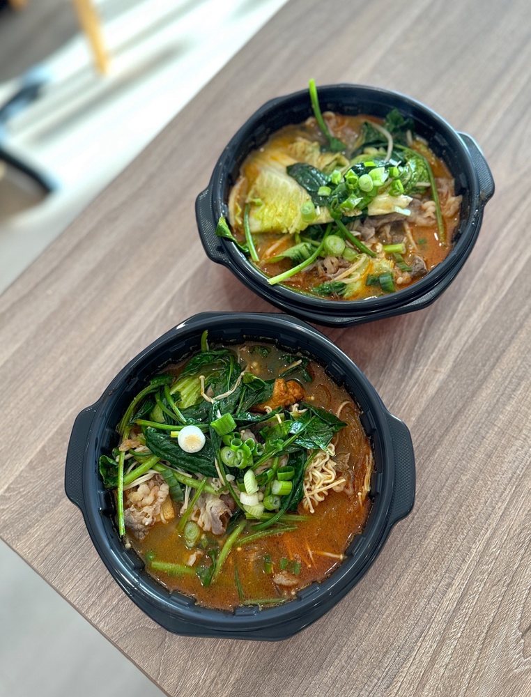 Two steaming bowls of X-Malatang loaded with leafy greens, noodles, and beef in a rich, spicy broth
